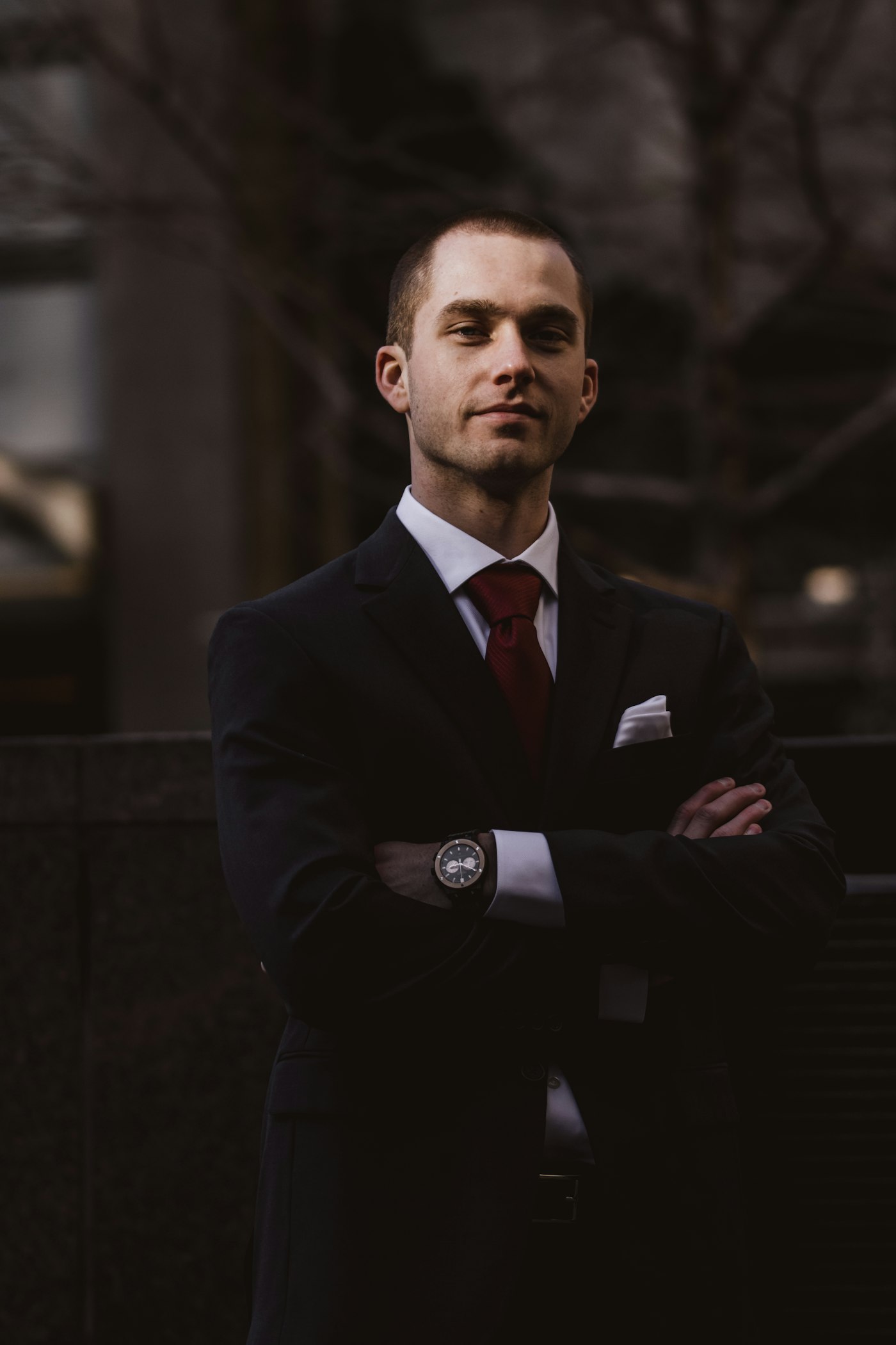 Man in a dark suit standing still with arms crossed against a dark urban background