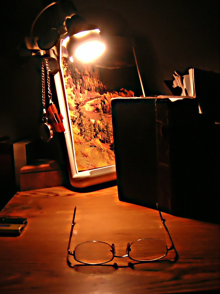 A lamp-lit writer's desk at night with glasses and an open book