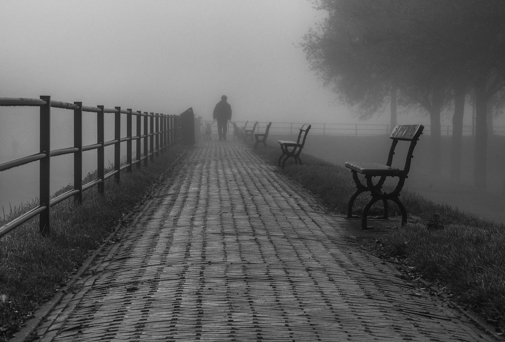 Man walking alone along a foggy riverside promenade in black and white