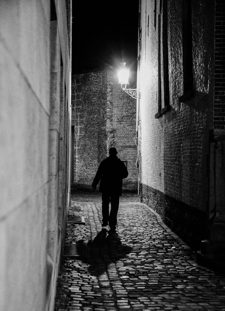 Man walking down a cobblestone alley at night under a street lamp, black and white