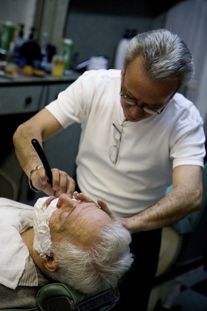 A barber giving a client a straight razor shave in a dim barbershop