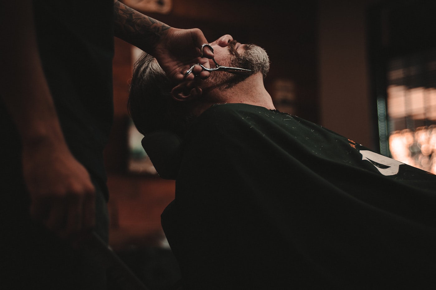 A barber trimming a client's beard in a dark barbershop