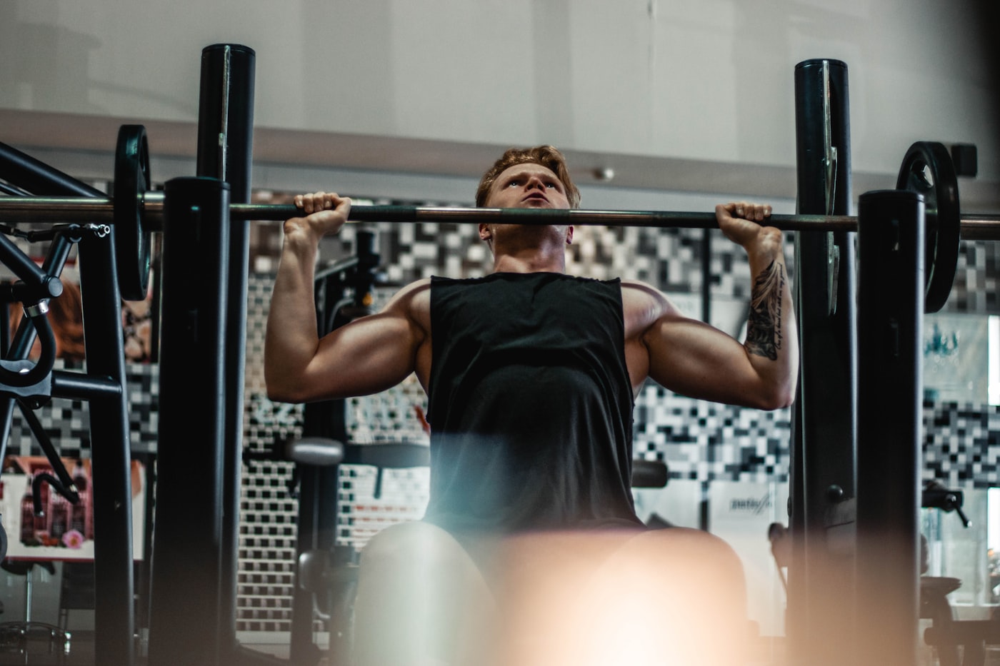 A man pressing a barbell overhead in a dim gym, seen from behind