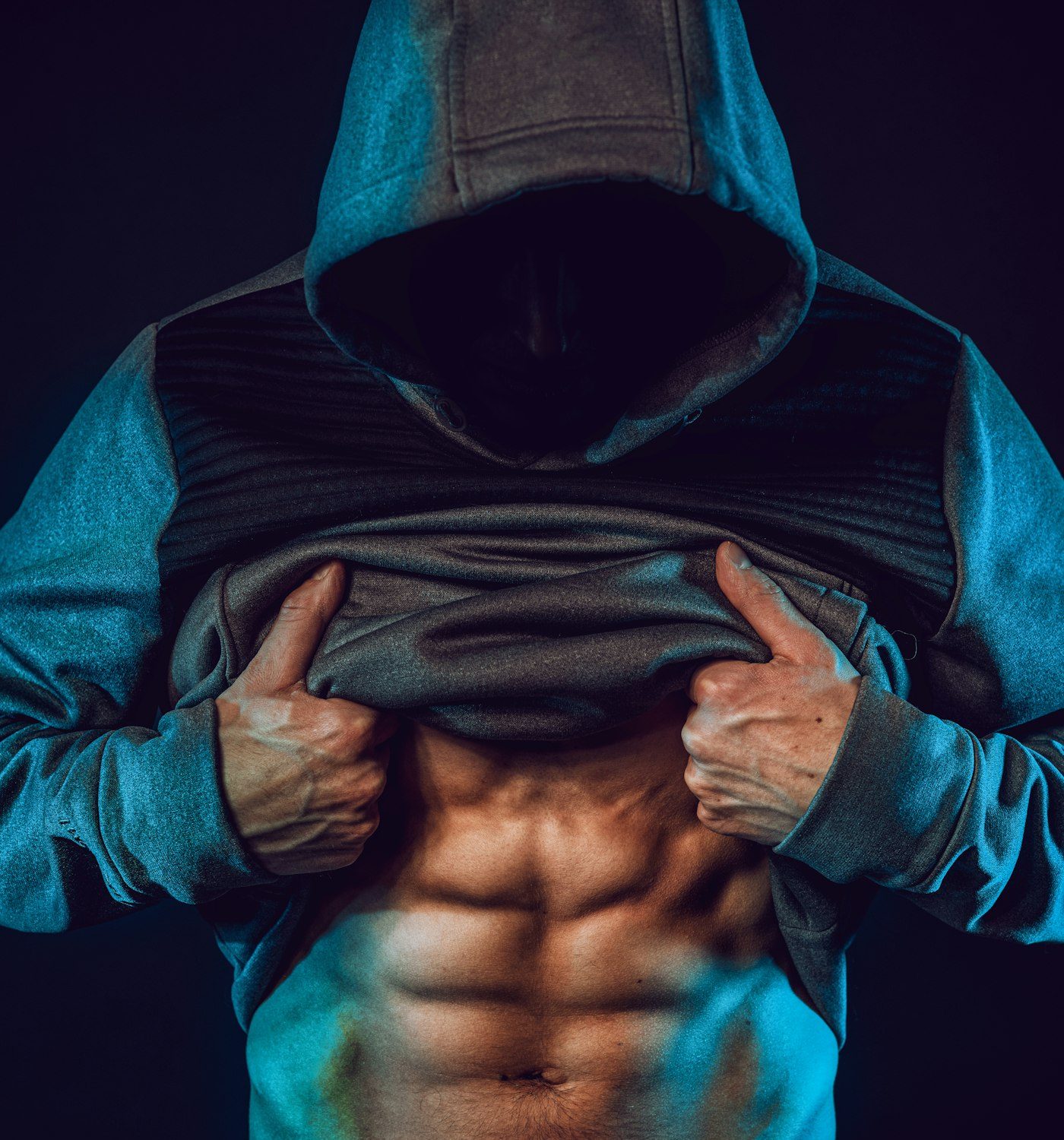 A hooded man lifting his sweatshirt to show a lean midsection in dark teal light