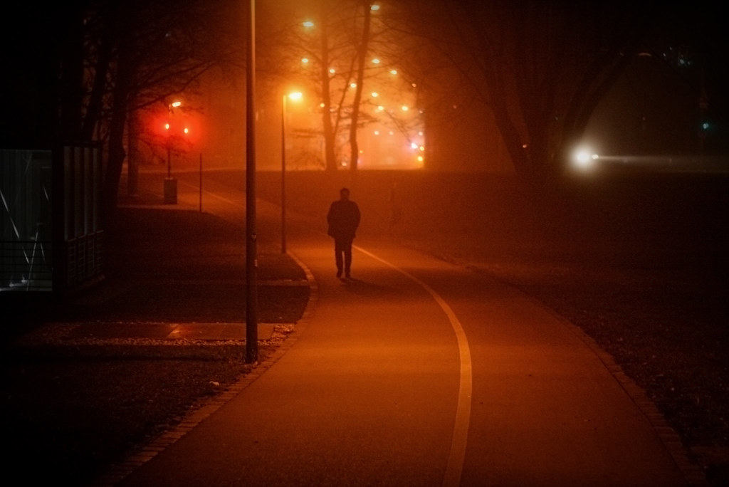 A lone figure walking down a foggy path at night under orange streetlights